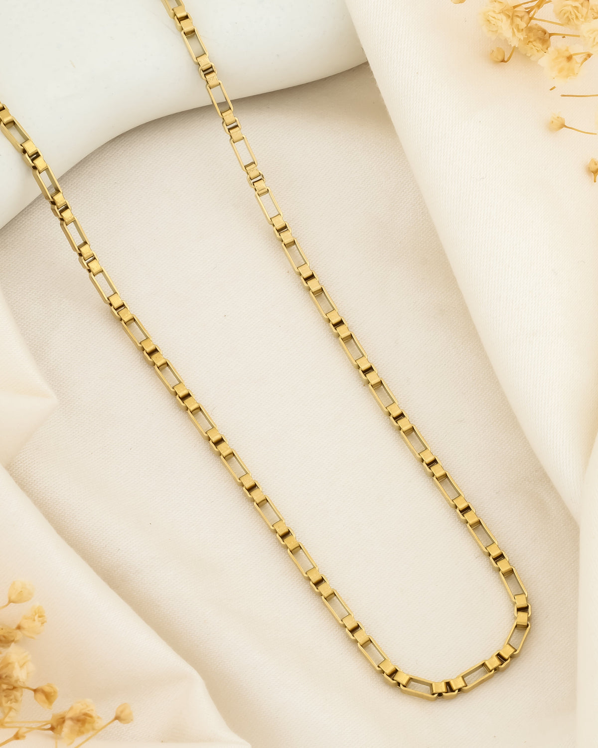 A gold chain necklace laid out on a white fabric surface with some dried flowers to the side.