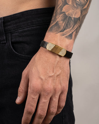 Hand wearing a gold and black bracelet with a tattooed arm against a neutral background